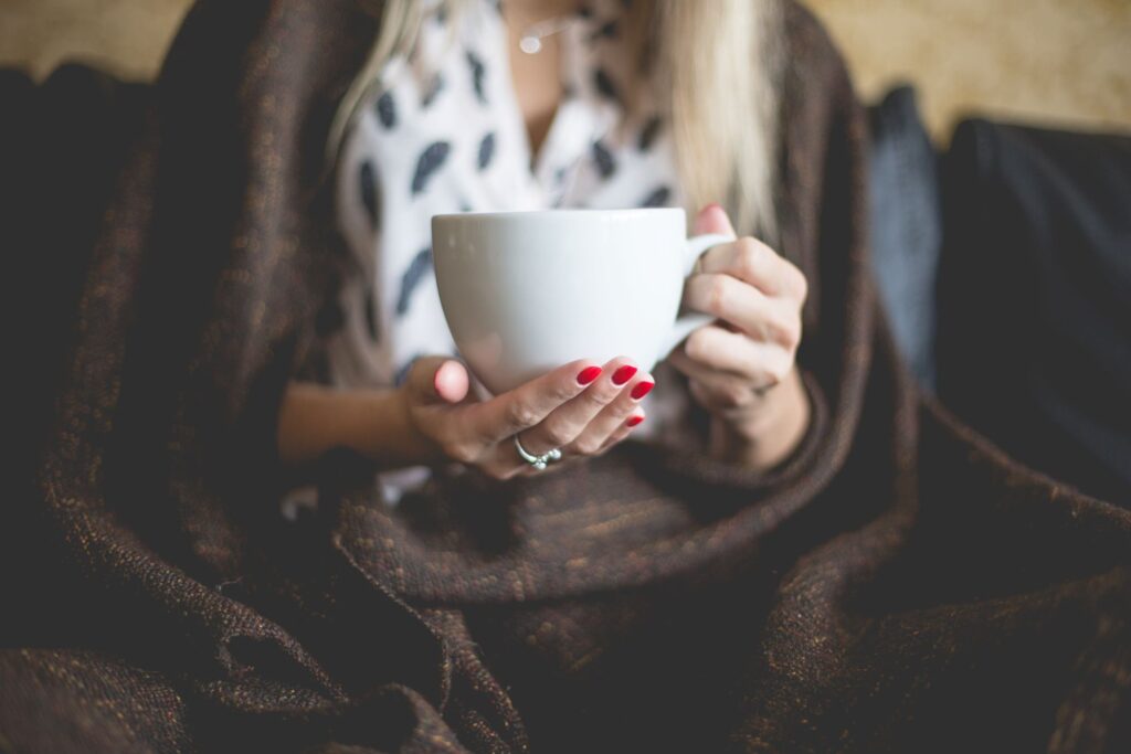 Woman wrapped in brown blanket  and with bright red nails holding a white coffee mug 
