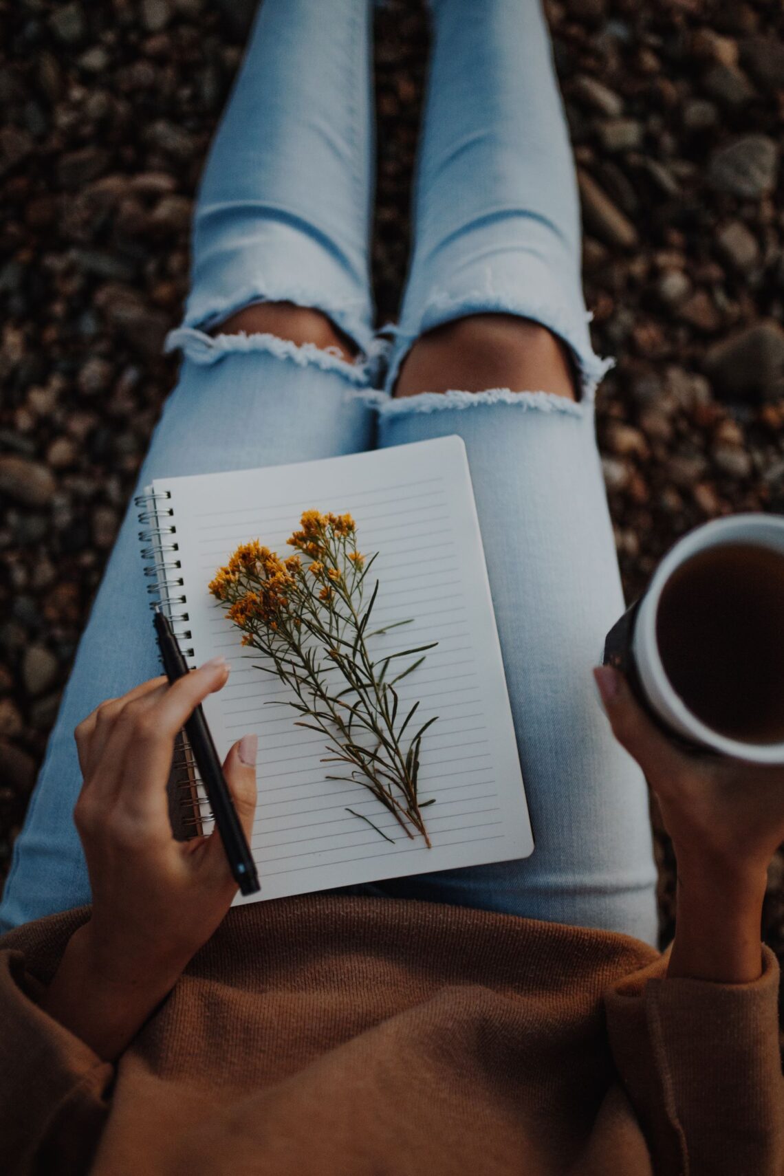 Woman sitting on the ground holding a mug of coffee and a journal with a yellow flower on it