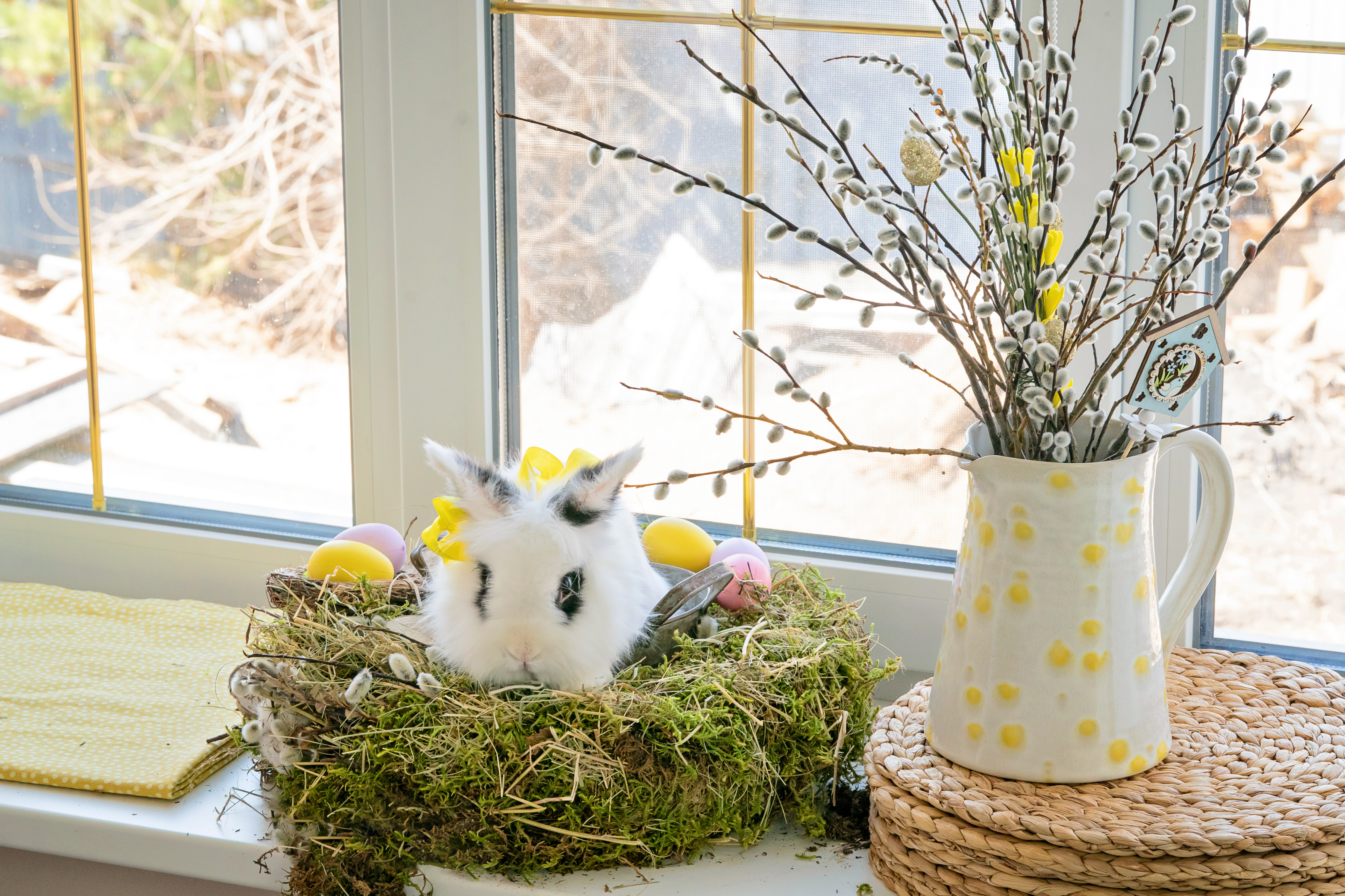 White rabbit sitting on green nest with eggs in it and a vase of flowers nearby